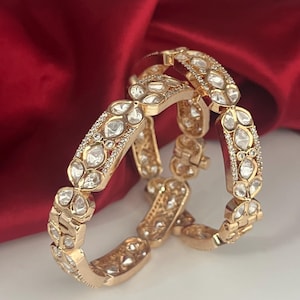 Peut inclure: Deux bracelets dorés ornés de pierres claires en forme de goutte et de petites pierres claires. Les bracelets sont posés sur un fond de satin rouge. Les bijoux ont un design orné.