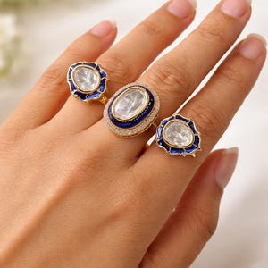 Puede incluir: Se muestran tres anillos dorados con piedras claras de forma ovalada. Los anillos tienen un borde de esmalte azul. El anillo central está rodeado de pequeñas piedras claras. Son joyas.