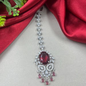 May include: A silver-toned headpiece with a large, oval, ruby-red gemstone at the center. The headpiece features a series of smaller, clear gemstones and teardrop-shaped red gems that dangle below. The background is a deep red fabric.
