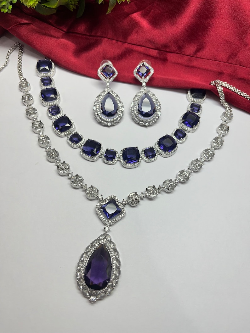 Peut inclure: Un ensemble de collier en argent avec un grand pendentif en forme de larme et deux plus petits pendentifs carr&eacute;s, tous sertis de pierres pr&eacute;cieuses violettes. L'ensemble comprend &eacute;galement une paire de boucles d'oreilles assorties avec des pierres pr&eacute;cieuses violettes en forme de larme.