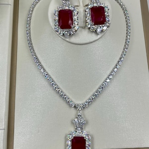 Pode incluir: Um conjunto de joias composto por um colar e brincos a condizer. O colar apresenta uma linha contínua de pedras transparentes com um grande pendente retangular de pedra vermelha escura. Os brincos espelham este design com pedras vermelhas.