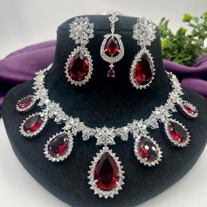 Peut inclure: Un ensemble de bijoux éblouissant comprenant un collier, des boucles d'oreilles et un bijou de tête. L'ensemble présente de grandes pierres précieuses rouges rubis en forme de goutte d'eau, chacune entourée d'un halo de pierres blanches scintillantes.