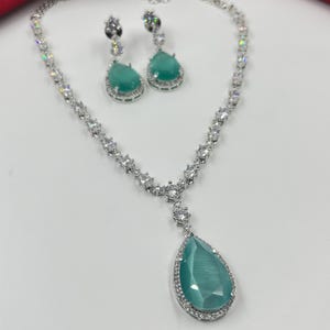 Peut inclure: Un collier en argent avec un pendentif en forme de larme orné d'une pierre précieuse verte entourée de petites pierres claires. Boucles d'oreilles assorties avec des pierres précieuses vertes et des pierres claires.