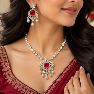 May include: A gold-toned necklace and matching earring set featuring square-cut red stones surrounded by clear white crystals and small white pearls. The necklace has a central pendant with a red stone, clear crystals, and dangling pearls.