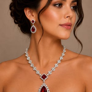 Puede incluir: Un deslumbrante conjunto de joyas con collar y pendientes. El collar tiene un gran colgante rojo en forma de lágrima, una gema roja cuadrada y una serie de piedras redondas y transparentes. Los pendientes reflejan el diseño del collar.