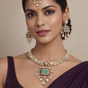 Puede incluir: Un conjunto de joyas dorado que incluye un collar, pendientes y una tiara. El conjunto presenta piedras de color esmeralda, detalles rojos y colgantes transparentes en forma de lágrima. El collar y los pendientes tienen un diseño a juego, y la tiara se lleva en la frente.