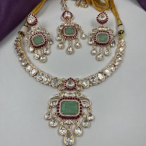 May include: A gold-toned jewelry set featuring a necklace, earrings, and a headpiece. The set includes a necklace with a large green stone pendant, earrings, and a headpiece, all with clear and red accents. The jewelry is set against a white background.