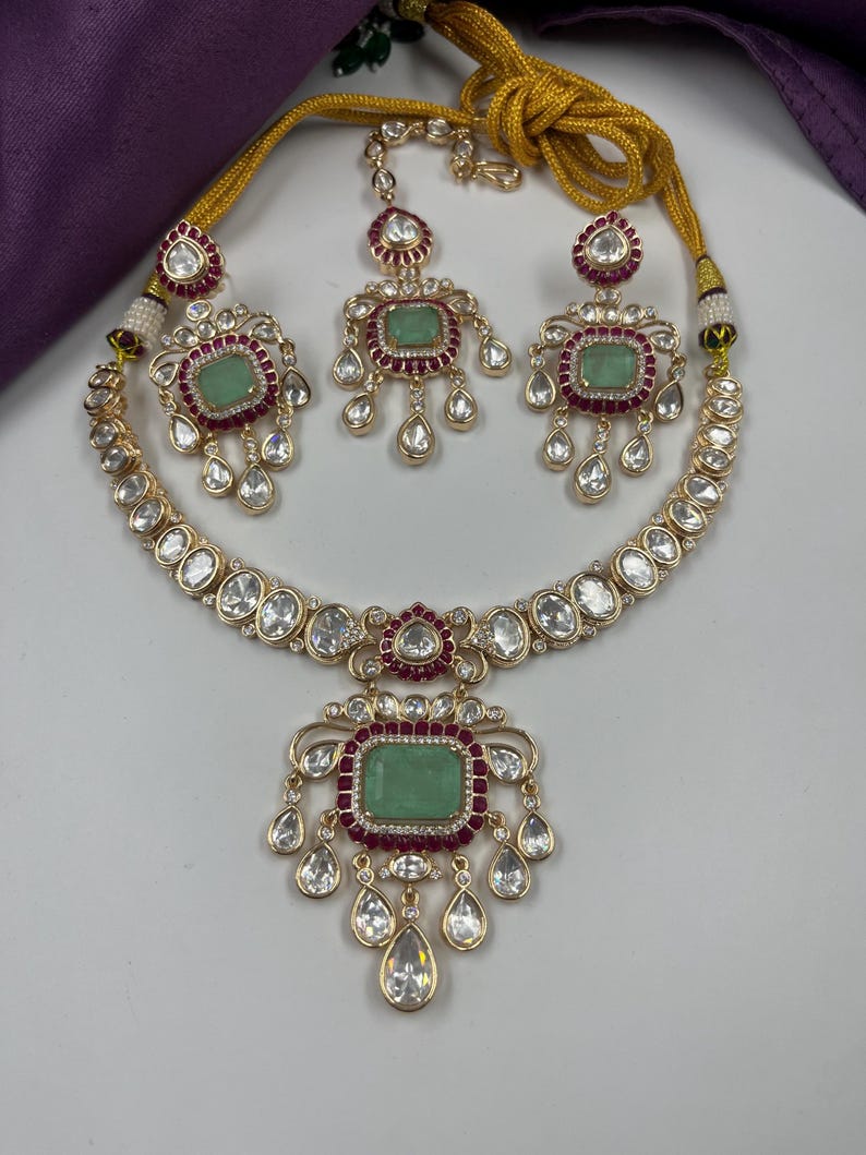 May include: A gold-toned jewelry set featuring a necklace, earrings, and a headpiece. The set includes a necklace with a large green stone pendant, earrings, and a headpiece, all with clear and red accents. The jewelry is set against a white background.