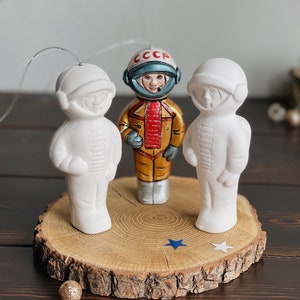 May include: Three white ceramic astronaut ornaments with a painted astronaut in the center. The painted astronaut is wearing a gold and red spacesuit with a helmet that says "CCCP". The ornaments are on a wooden slice with blue and white stars.