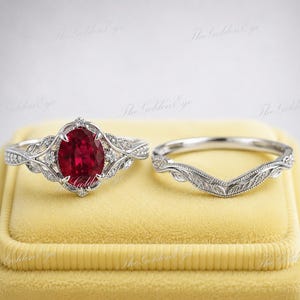 May include: A silver-toned ring set featuring an oval-cut deep red gemstone center stone surrounded by small clear stones on a band with intricate leaf-like engravings, paired with a matching curved band featuring similar leaf-like metal detailing.