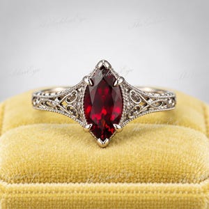 May include: A ring featuring a marquise-cut deep red garnet gemstone set in a silver-toned metal band. The band displays intricate filigree scrollwork and milgrain detailing along the shoulders, framing the central stone with a prong setting.
