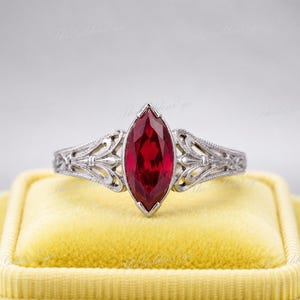 May include: A silver-toned ring featuring a central marquise-cut deep red gemstone. The metal band displays intricate openwork filigree detailing along the sides, extending from the stone setting to the shank, creating a delicate and ornate appearance.