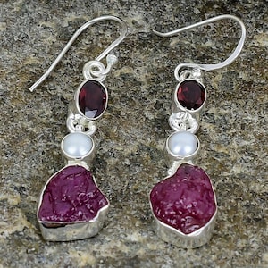 May include: Silver dangle earrings featuring a deep red, oval-cut gemstone, a small white pearl, and a large, irregular-shaped, deep pink gemstone. The earrings are displayed on a textured gray stone surface.