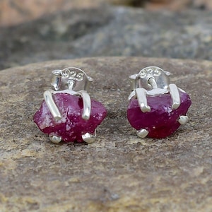 Può includere: Un paio di orecchini in argento sterling con pietre preziose di rubino grezzo. Gli orecchini sono incastonati in uno stile semplice e minimalista, con i rubini tenuti in posizione da delicate griffe. I rubini sono di un colore rosso intenso e hanno una finitura naturale non lucidata.