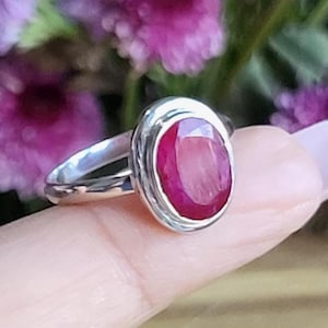 May include: A silver ring with an oval, deep red gemstone. The ring is held up against a background of purple flowers. The gemstone is the focal point, showcasing its rich color and polished surface.