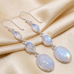 Pode incluir: Brincos pendurados em prata com três pedras da lua ovais e em forma de gota. Os brincos, com fechos de gancho, são exibidos em cetim. As pedras da lua têm um brilho iridescente. Comprimento: aproximadamente 6 cm.