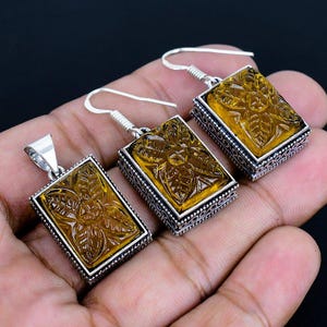 May include: A jewelry set featuring a pendant and two earrings. Each piece has a rectangular, amber-colored stone with a carved floral design, set in a silver-colored metal frame. The earrings have hook closures.