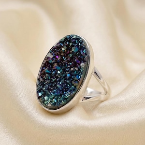 Puede incluir: Anillo de plata con una piedra drusa ovalada. La piedra tiene una superficie brillante e iridiscente con tonos azules, morados y verdes. El anillo tiene un diseño sencillo y elegante, perfecto para el uso diario.