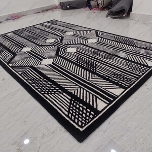 Może przedstawiać: Czarno-biały dywan w geometryczne wzory o nowoczesnym designie. Dywan ma białe tło z czarnymi liniami i kształtami.