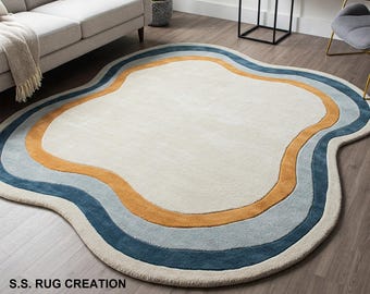 May include: A cream-colored, irregularly shaped rug with a wavy design. The rug features concentric bands of color, including blue, orange, and light blue, creating a layered effect. The rug is placed on a hardwood floor.