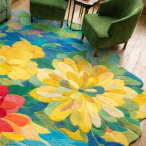May include: A large, floral-shaped rug with a vibrant design. The rug features large yellow, red, and blue flower patterns with green leaves. The rug is placed on a wooden floor, with green armchairs and a small table in the background.