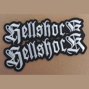 May include: Two black embroidered patches with the word "Hellshock" in a gothic font, outlined in white. The patches are on a tan surface. These patches are likely for clothing or accessories.