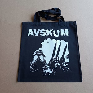 May include: Black tote bag with white print. The print features a soldier wearing a gas mask and holding a rifle. The text "AVSKUM" is printed in large white letters at the top of the bag.