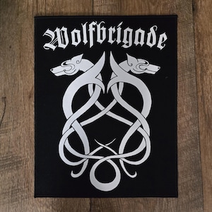 May include: Black rectangular patch with the word "Wolfbrigade" in white gothic font. The patch features a symmetrical design of two wolf heads and intertwined lines in white.