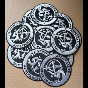 May include: A pile of black and white embroidered patches. Each patch is circular with the words "GRIND CORE" and "ANTI FASCIST" around the edge. A central graphic depicts a figure with a weapon. The patches are on a tan surface.