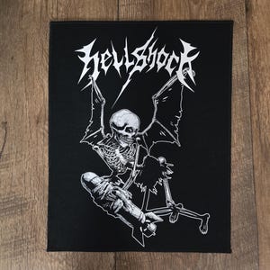 Puede incluir: Parche negro con una ilustración blanca de un esqueleto con alas de murciélago, sosteniendo una bomba, y la palabra "Hellshock" en una fuente estilizada. El diseño es una imagen gráfica audaz.