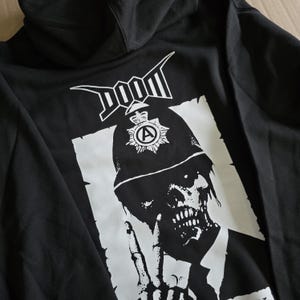 Könnte beinhalten: Schwarzer Kapuzenpullover mit einem weißen Grafikdruck eines Schädels, der einen Polizeihut trägt, mit dem Wort "DOOM" darüber.