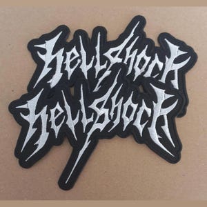Op de afbeelding: Zwarte geborduurde patches met de tekst "Hellshock" in een witte, gotische letter. De patches hebben een zwarte rand en zijn bedoeld voor kleding of accessoires.