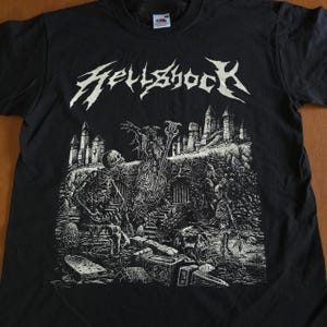 May include: Black t-shirt with the white text "Hellshock" above a detailed graphic. The graphic depicts a skeletal figure, tombstones, and a castle-like structure. The design is a detailed, black and white illustration.