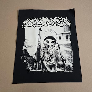 May include: Black and white patch featuring the word "Dystopia" in a stylized font above a graphic of a person holding a rifle. The person's face is partially covered. The patch is rectangular.