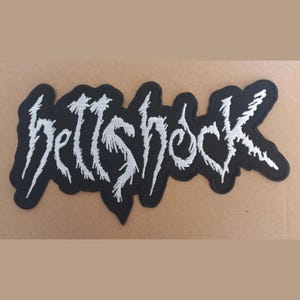 Pode incluir: Um patch preto com o texto branco "hellshock" em uma fonte estilizada e irregular. O patch tem uma borda preta e é projetado para ser costurado ou passado a ferro em roupas ou acessórios.