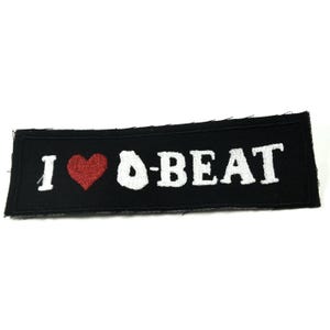 Op de afbeelding: Zwarte geborduurde patch met witte tekst die "I ♥ O-BEAT" luidt.