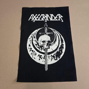 Puede incluir: Parche bordado en blanco y negro con el nombre de la banda "AXGRINDER" en una fuente estilizada. El parche presenta una calavera con una espada que la atraviesa, rodeada de un círculo de runas.