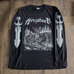 May include: Black long-sleeve shirt with a graphic design. The shirt features the word "Hellshock" in a stylized font, along with a detailed black and white illustration of a graveyard scene. Large sword graphics are printed on the sleeves.