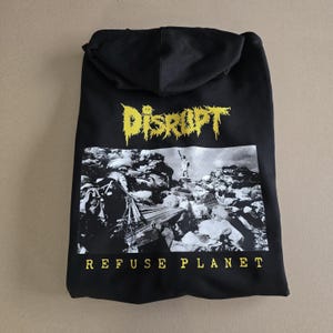 May include: Black hooded sweatshirt with the word "DISRUPT" in yellow, jagged lettering above a black and white graphic of a statue on a pile of debris. The words "REFUSE PLANET" are printed in yellow below the graphic.