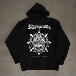 Peut inclure: Sweat à capuche noir avec un motif blanc au dos. Le motif représente un crâne, une croix et d'autres éléments, avec le texte "SKITSYSTEM" au-dessus et "DÖDEN ÄR MIN MASKINIST" en dessous.