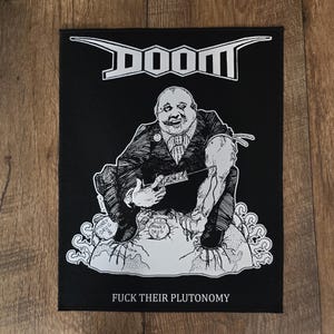 Puede incluir: Parche negro con la palabra "DOOM" en plata en la parte superior. Debajo, una ilustración en blanco y negro de un hombre con traje sosteniendo un cuchillo, sentado sobre sacos de dinero. El texto "FUCK THEIR PLUTONOMY" está en la parte inferior.