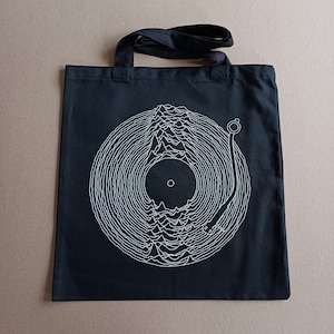 Könnte beinhalten: Schwarze Tragetasche mit einer weißen Grafik eines Schallplatten mit einem stilisierten Wellenmuster. Die Schallplatte befindet sich auf einem Plattenspieler mit einer Nadel.