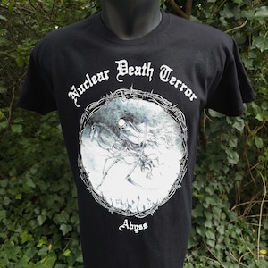May include: Black t-shirt with the words "Nuclear Death Terror" in white script above a circular graphic. The graphic depicts a skeletal figure on horseback with a scythe, surrounded by barbed wire. The word "Abyss" is below the graphic.
