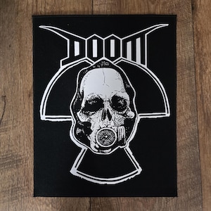 May include: Black patch featuring a white skull wearing a gas mask, with the word "DOOM" above. The design is a stylized, graphic illustration. The patch is rectangular and appears to be made of fabric.