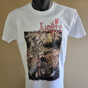 Op de afbeelding: Wit T-shirt met een grafisch ontwerp. Het ontwerp toont het woord "Amerix" in rode, gestileerde letters en een skeletfiguur in een donkere omgeving. Het woord "Monolith" staat onderaan het ontwerp.
