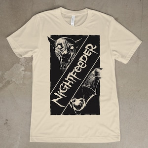 Op de afbeelding: Crèmekleurig T-shirt met een zwart grafisch ontwerp. Het ontwerp bevat een gestileerde illustratie van een vleermuis en een wezen met gloeiende ogen, samen met het woord "NIGHTFEEDER" in een diagonale lettertype.