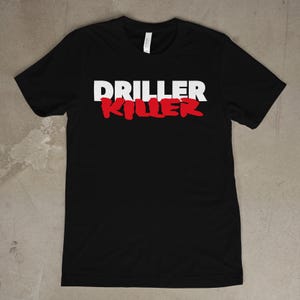 Op de afbeelding: Zwart t-shirt met de tekst "DRILLER KILLER" op de voorkant. Het woord "DRILLER" is wit en "KILLER" rood. Het shirt is gemaakt van een zacht materiaal.