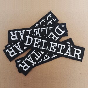 Könnte beinhalten: Ein Stapel schwarzer Stoffpatches mit weiß gestickten Buchstaben, die "DELETAR" lesen.