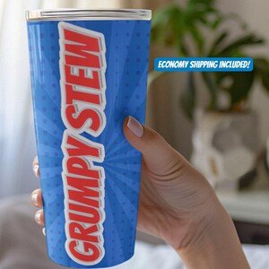 Puede incluir: Un vaso azul con las palabras "GRUMPY STEW" en letras rojas grandes. El vaso tiene un borde blanco y una tapa plateada. El fondo presenta un diseño de explosión radial azul con pequeños puntos blancos.