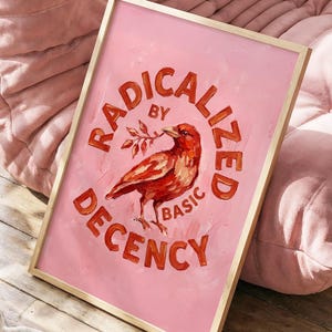 Peut inclure: Une affiche encadrée avec un fond rose et un cadre doré. L'impression présente un oiseau rouge tenant une branche, avec les mots "RADICALIZED BY BASIC DECENCY" en texte rouge. L'œuvre est exposée sur un sol en bois.